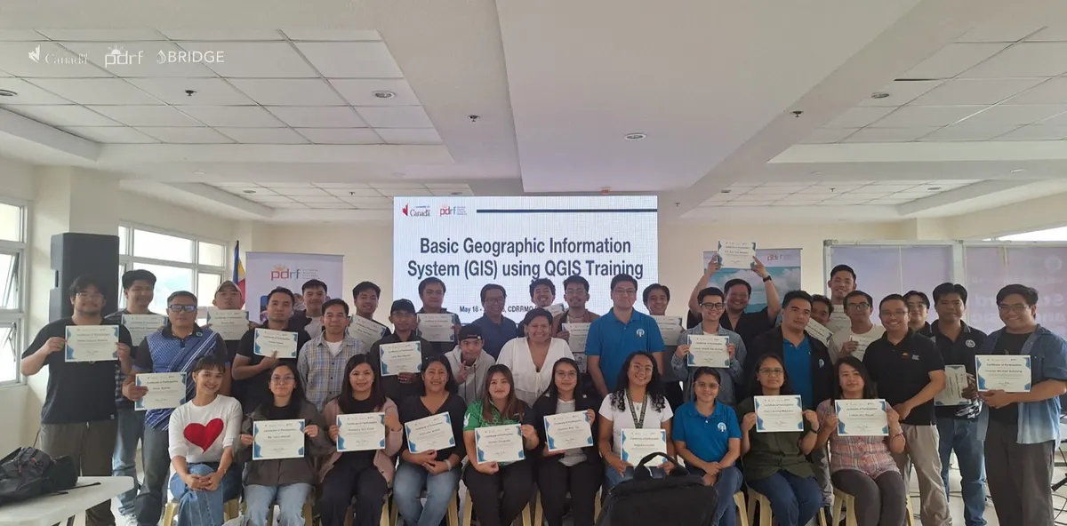Participants holding their certificates of participation in front of a screen reading “Basic Geographic Information System (GIS) using QGIS Training”.